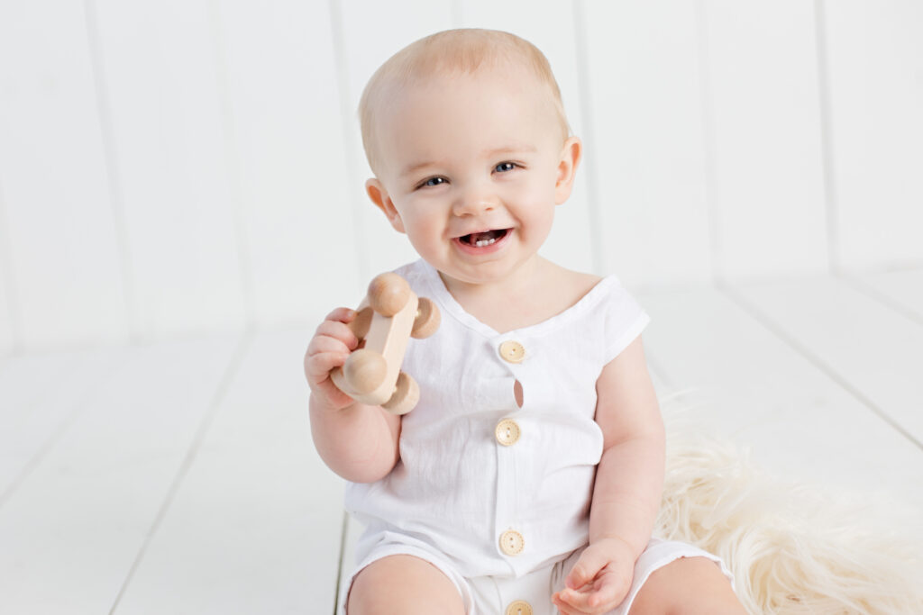 1 year old smiling baby boy in white romper holding wooden car toy with white wood background