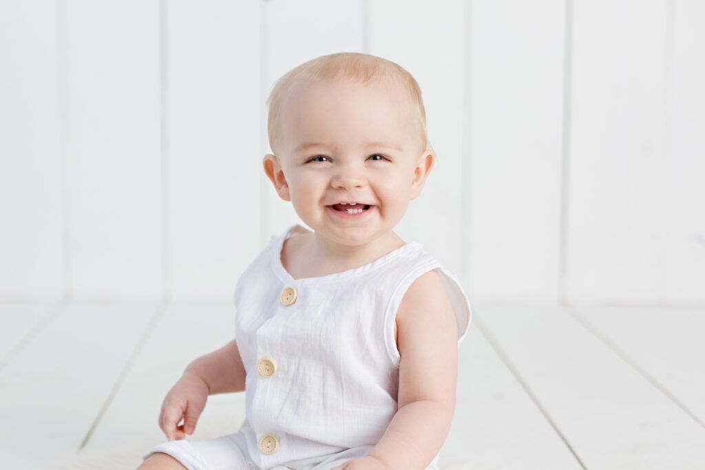 1 year old smiling baby boy in white romper with white wood background