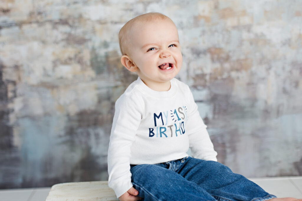 1 year old smiling baby boy in my first birthday shirt sitting on a wooden box
