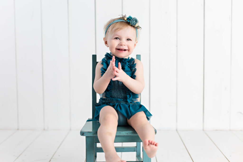 Baby girl sitter session with teal romper and garden tieback sitting on a teal chair with a white wood background.
