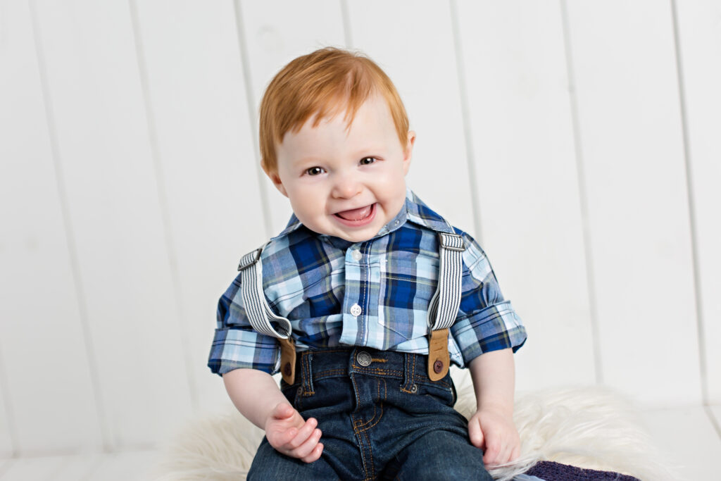 Baby boy sitter session with suspenders and blue plaid shirt on white wood background.