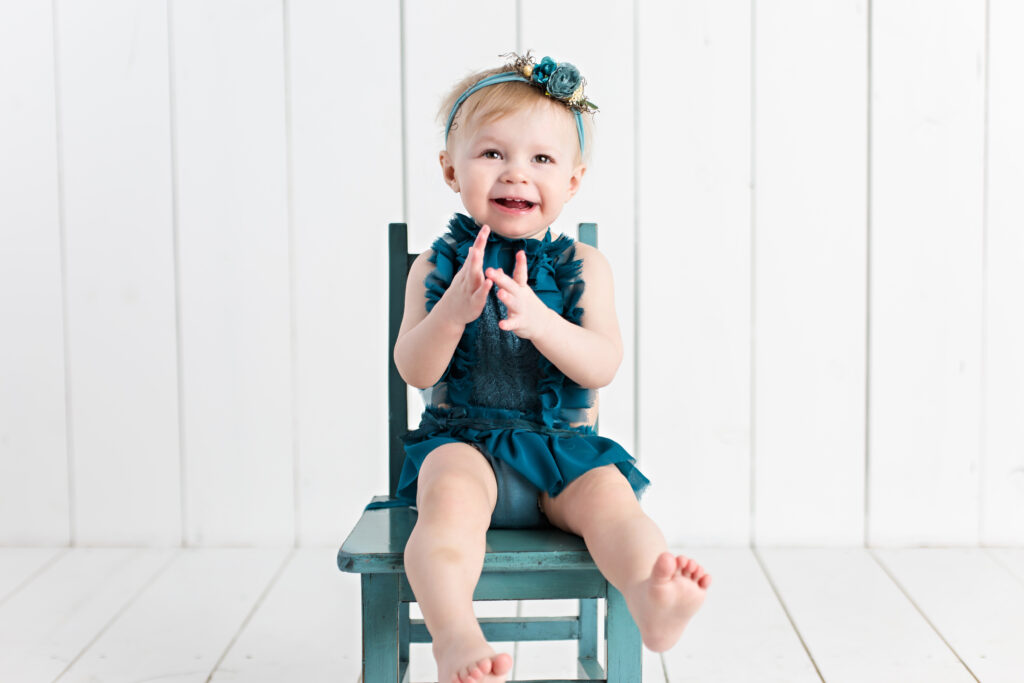 Baby girl sitter session with teal romper and garden tieback sitting on a teal chair with a white wood background.