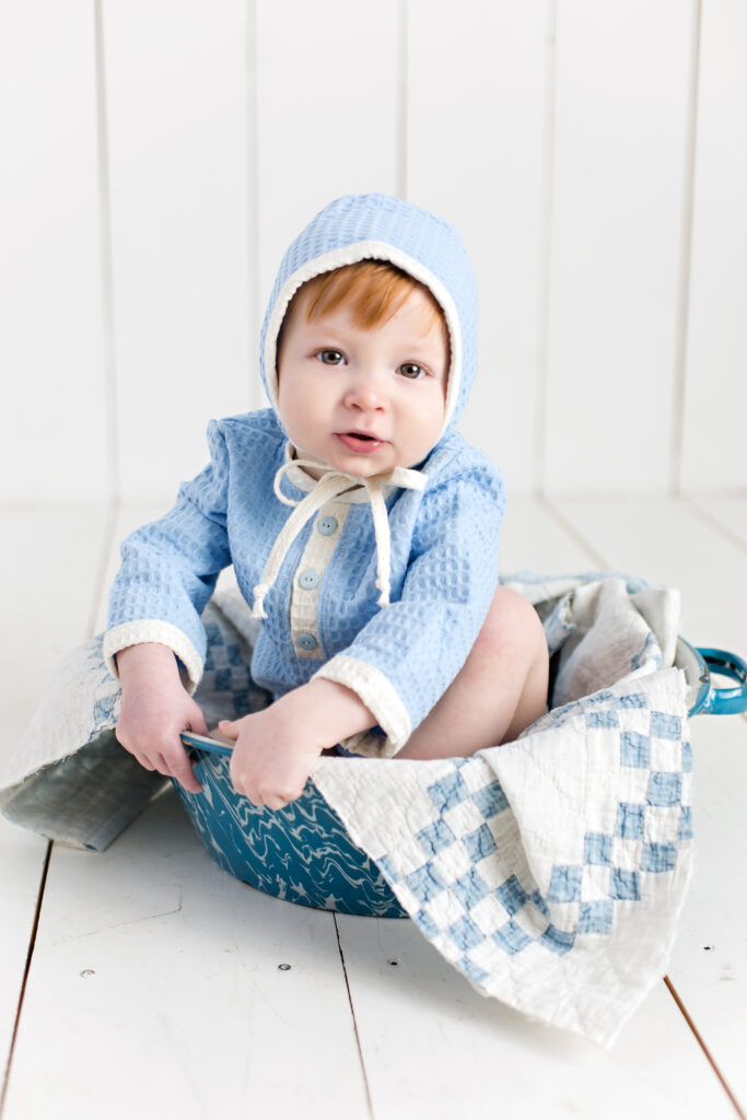 Baby boy sitter session with light blue romper and bonnet sitting in a bowl with a quilt piece.