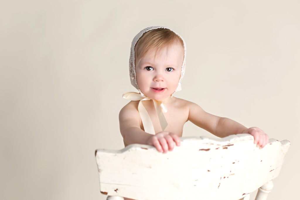 Baby girl sitter session with lace bonnet and cream seamless background and antique chair.