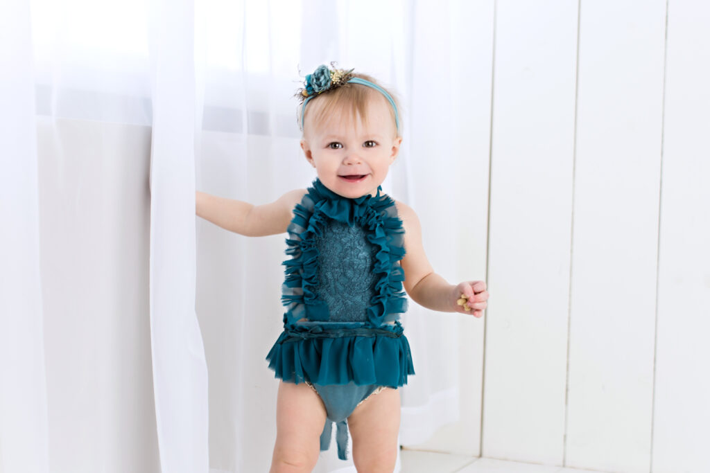 Baby girl sitter session with teal romper and garden tieback with flowing white curtain and white wood background.