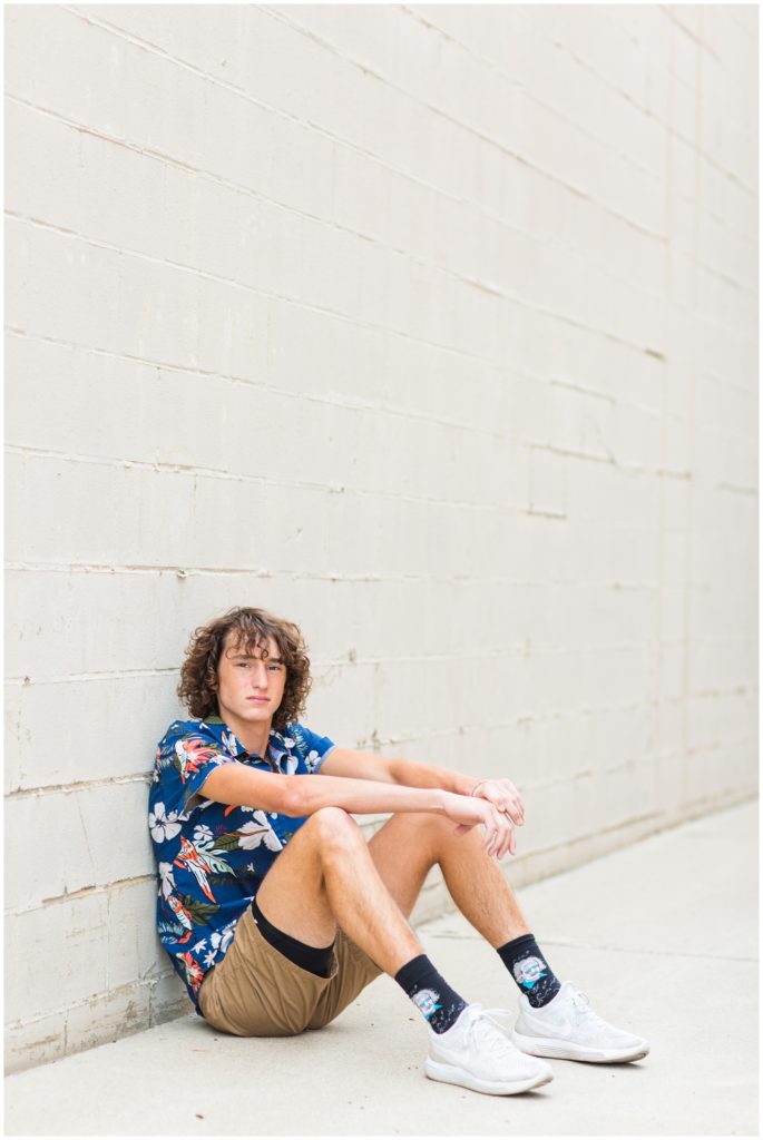 Senior boy poses sitting by a white wall in downtown Algona, IA CB Studio Photography