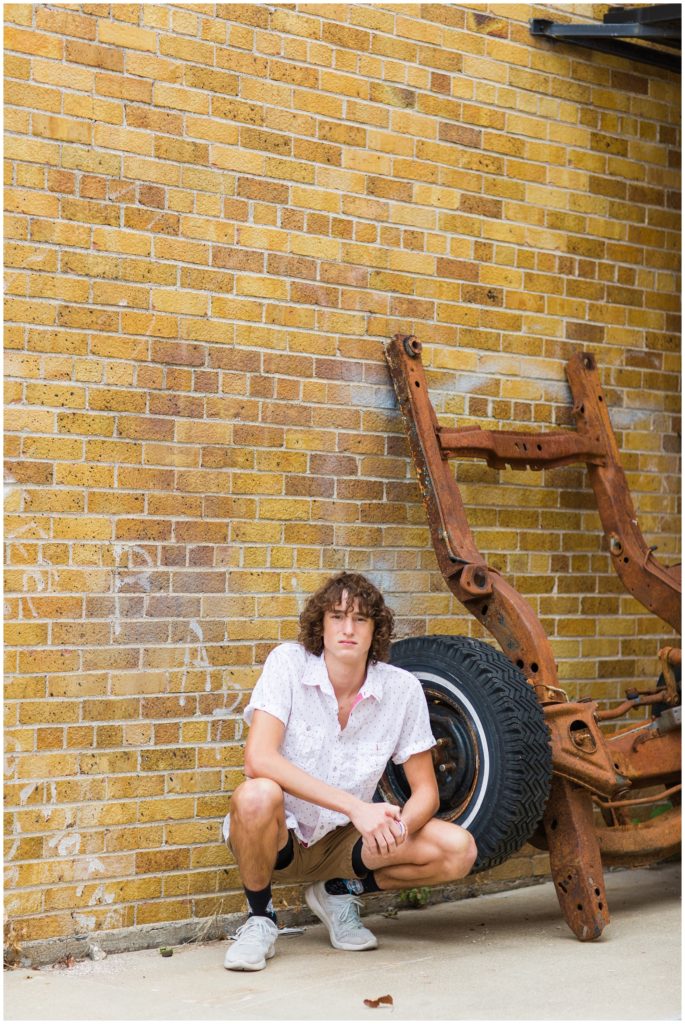 Senior boy poses squatting by an old rusty wheel base and brick wall in downtown Algona, IA CB Studio Photography