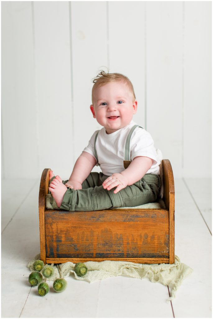 6 month old baby boy sitting in a small wooden baby bed wearing suspenders with a little bit of greenery in the decor | Sitter Session | Iowa Baby Photographer | CB Studio