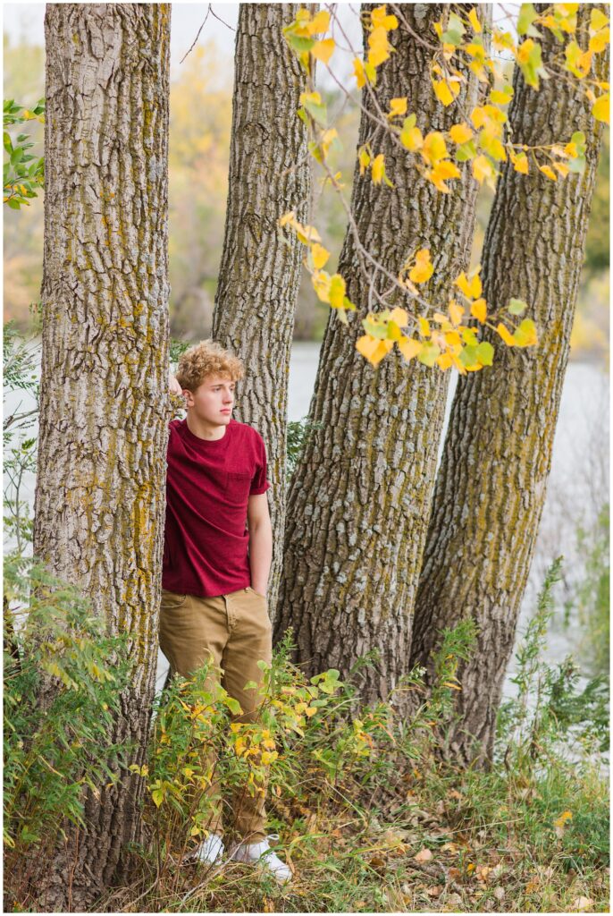 Senior boy leaning on a tree surrounded by fall foliage with a lake in the background at Plum Creek Wildlife Area | Iowa Senior Photographer | CB Studio