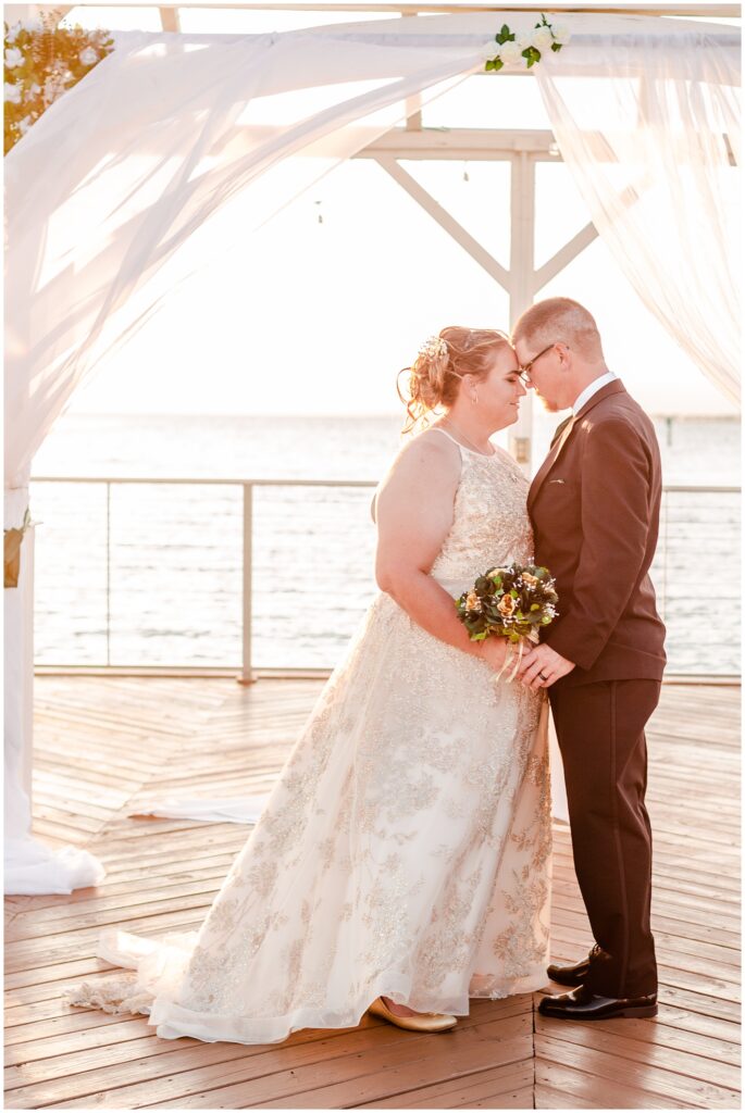 Bride and groom romantically standing under a white wedding pergola with white drapes flowing in the breeze on the Godfrey Hotel bay pier over looking the bay at sunset | Tampa Bay Wedding | CB Studio