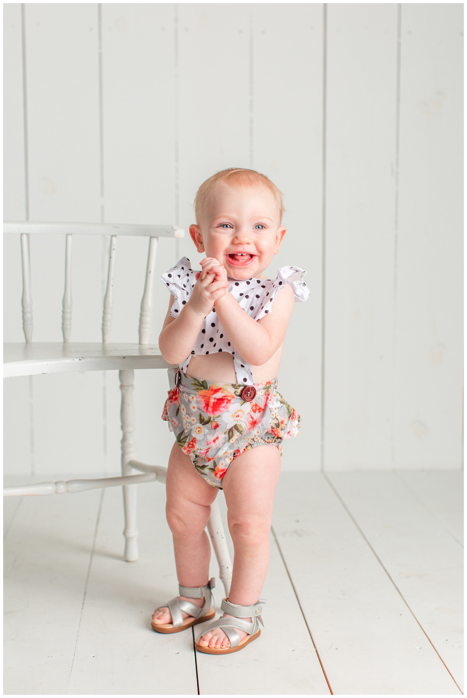 One year old baby girl standing on a white wood background wearing a floral and polka dot romper, smiling and clasping her hands | Iowa Baby Photographer | CB Studio