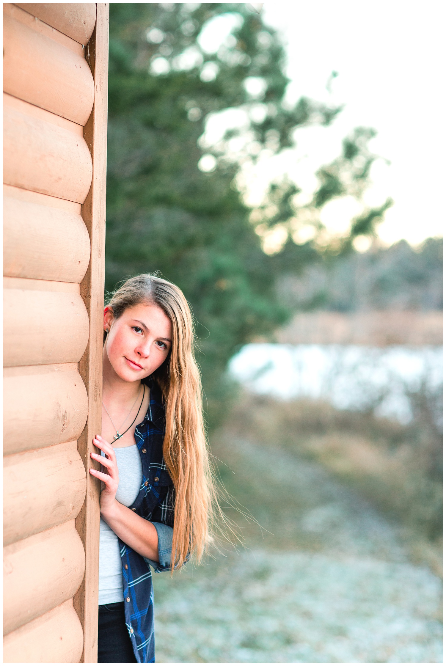 Senior girl peeking out from behind a wood building in a park with a lake during the fall | Iowa Senior Photographer | CB Studio