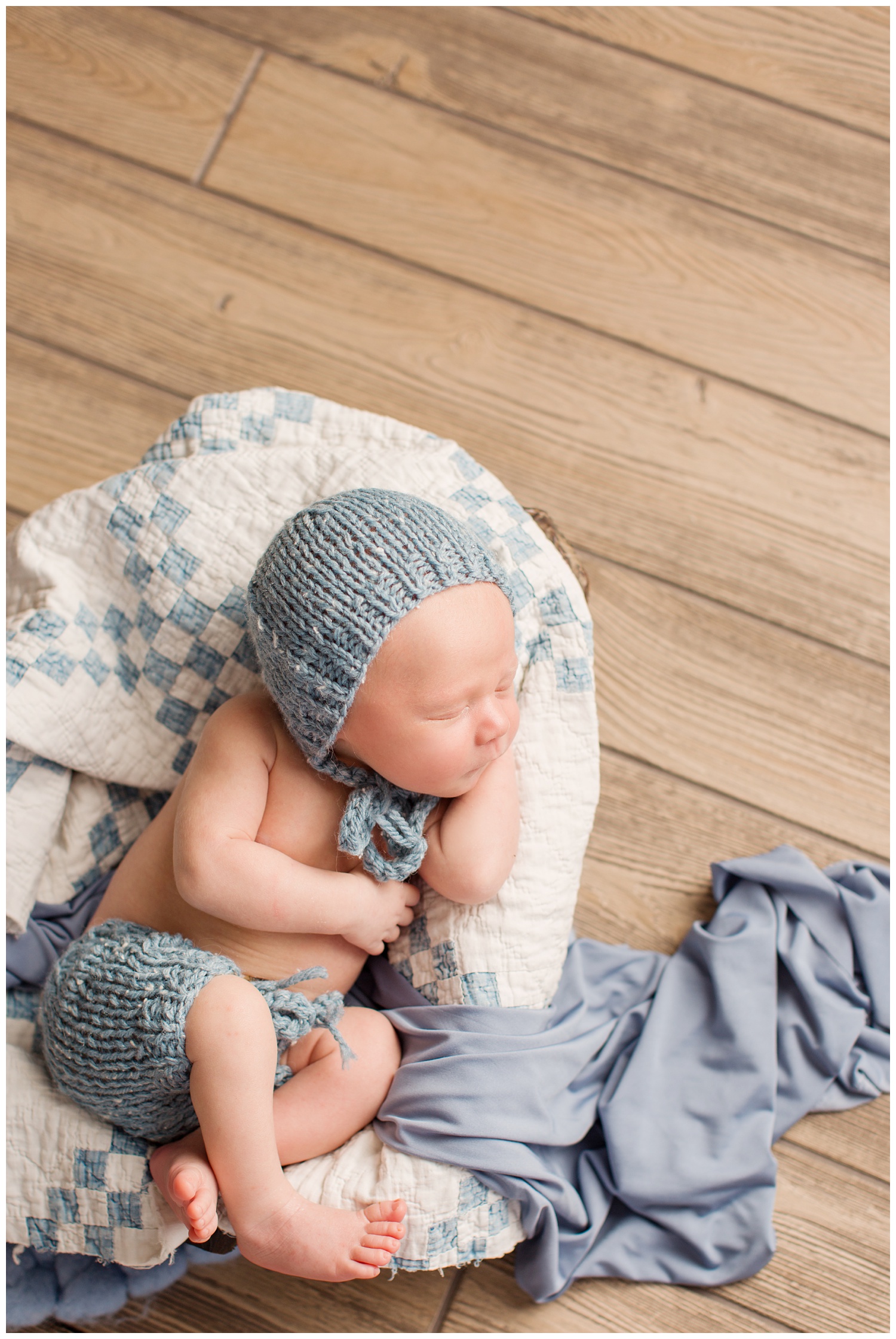 Newborn baby laying in a bowl covered with a quilt wearing a blue knit bonnet and shorts