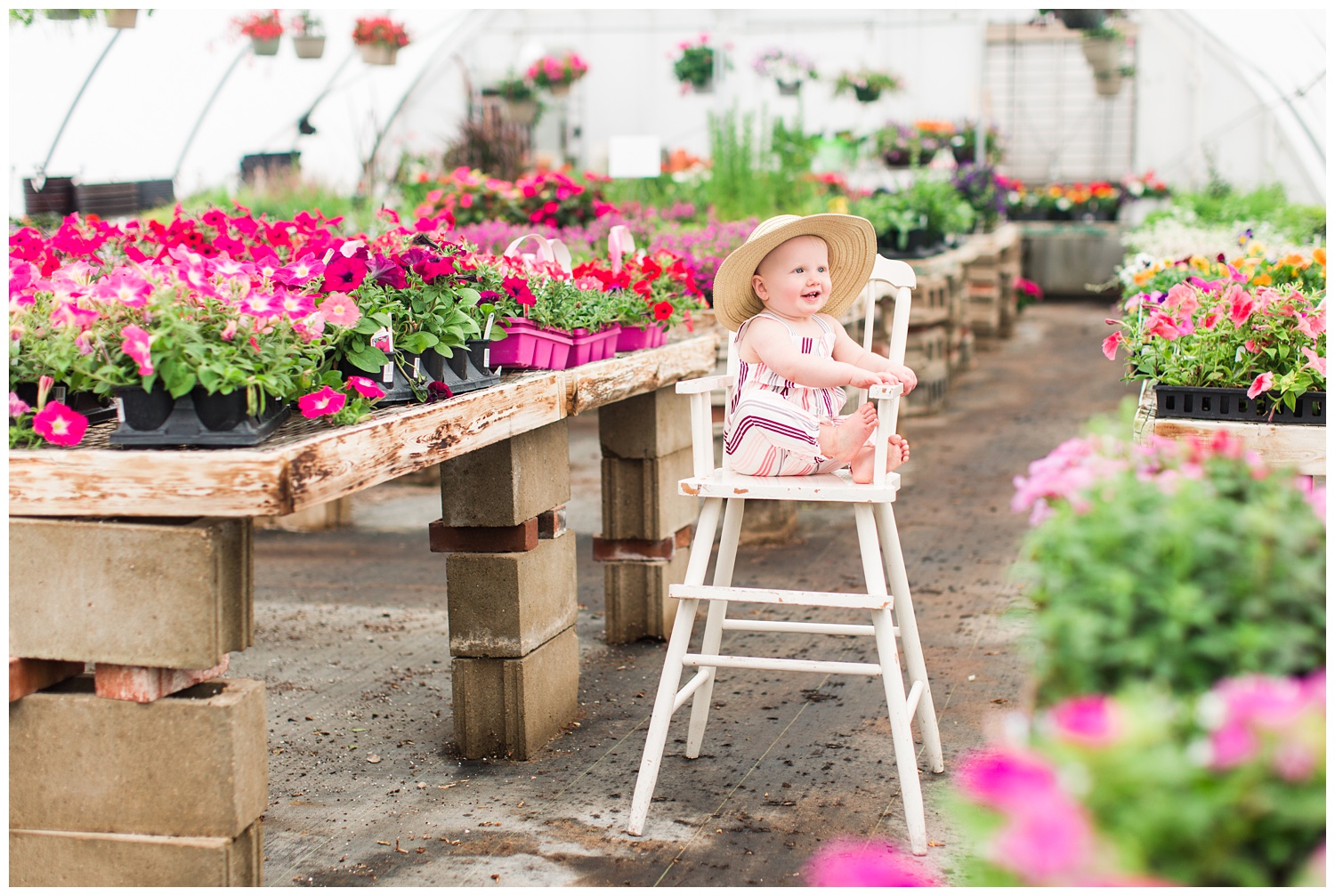 Baby Ivy sitting in a cream antique high chair surrounded by florals in a greenhouse wearing a fun floppy hat.
