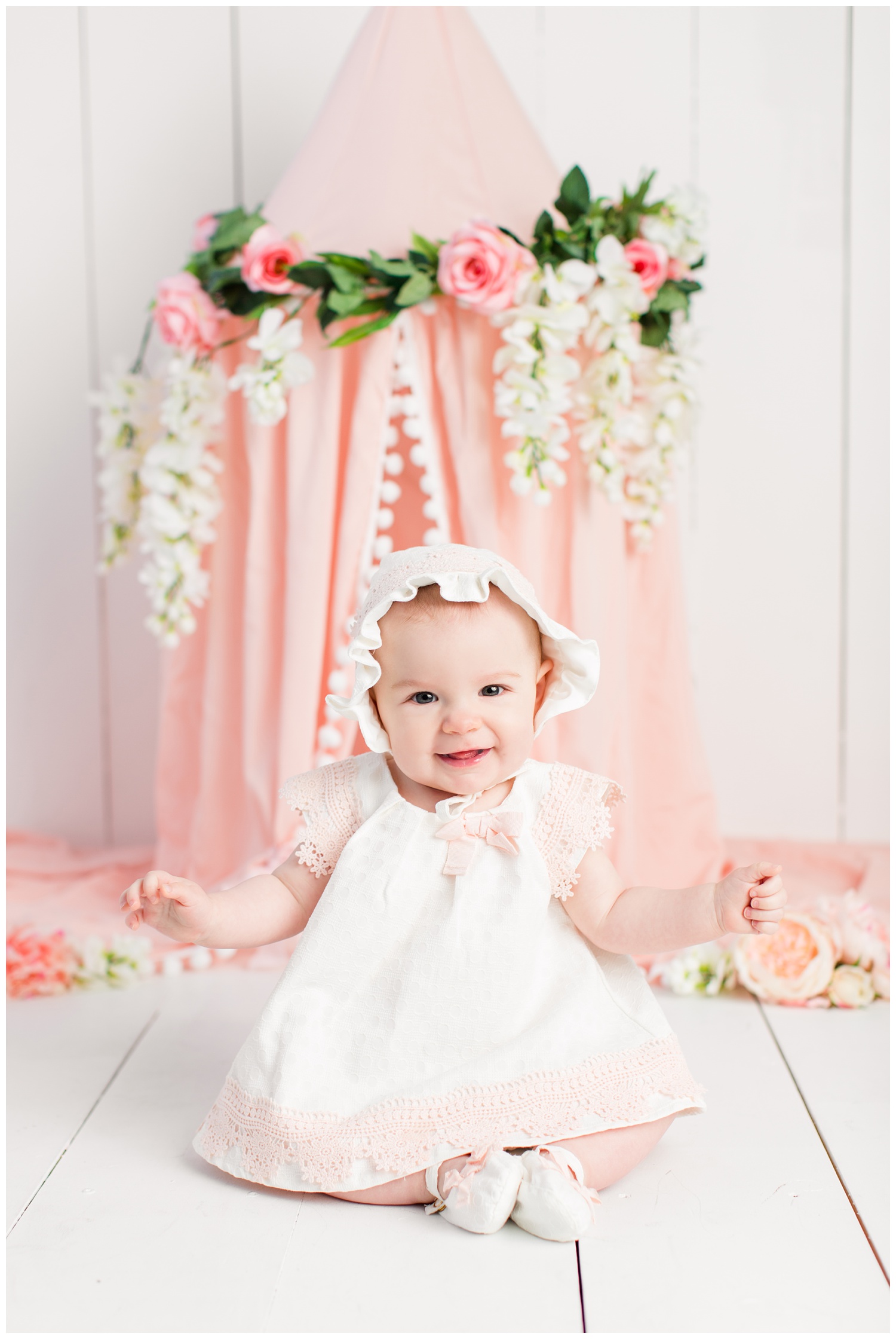 Baby Nora poses in a cream and pink babydoll dress and bonnet from TJ Maxx in front of a pink bed canopy decorated in florals.