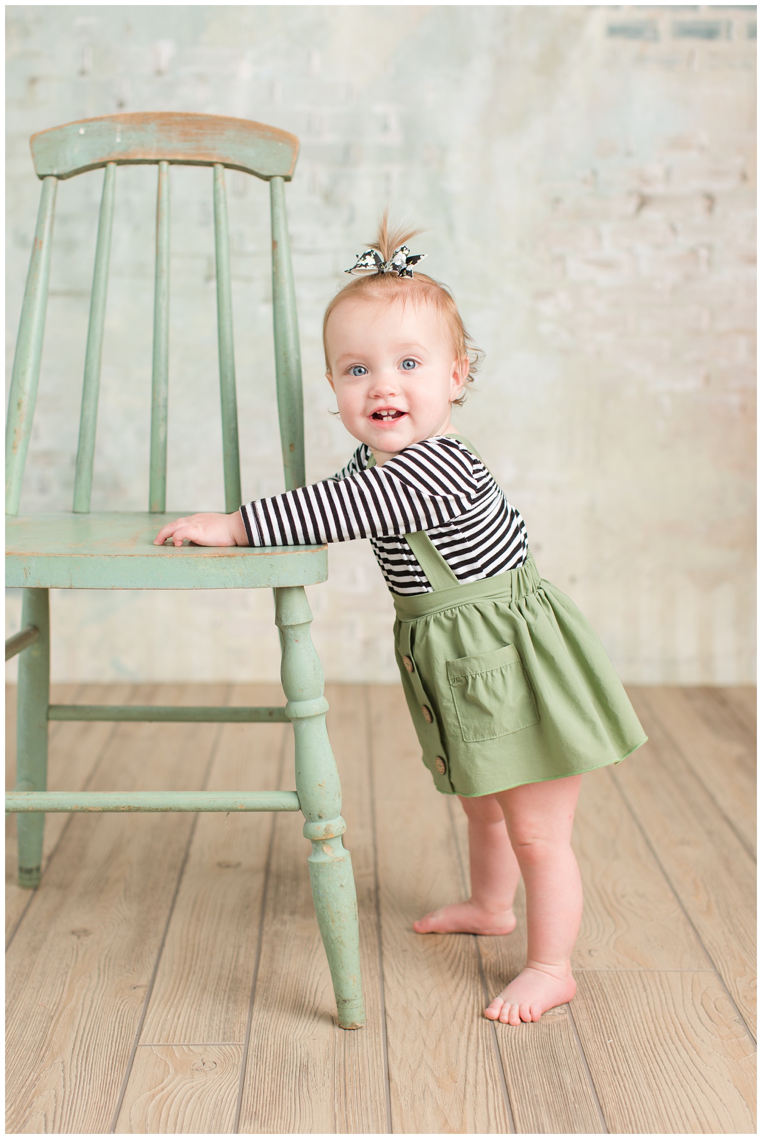 Baby Cara stands next to a rustic sage green chair wearing an sage green skirt and black and white striped shirt | CB Studio