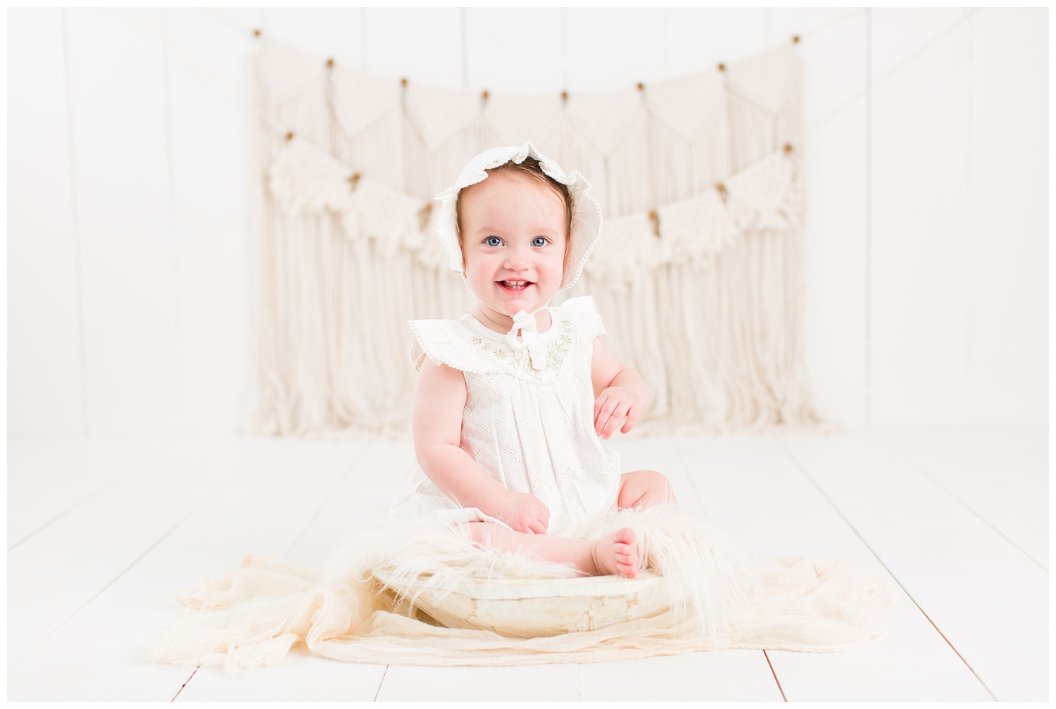 Baby Cara dressed in a vintage white romper and bonnet sitting in a rustic cream bowl with a boho macrame background | CB Studio