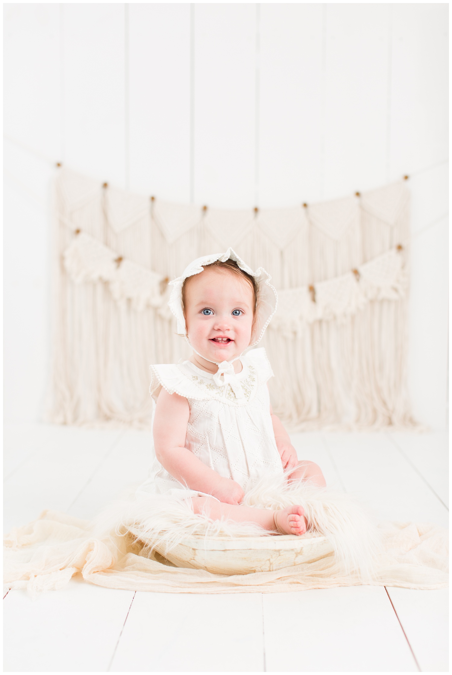 Baby Cara dressed in a vintage white romper and bonnet sitting in a rustic cream bowl with a boho macrame background | CB Studio