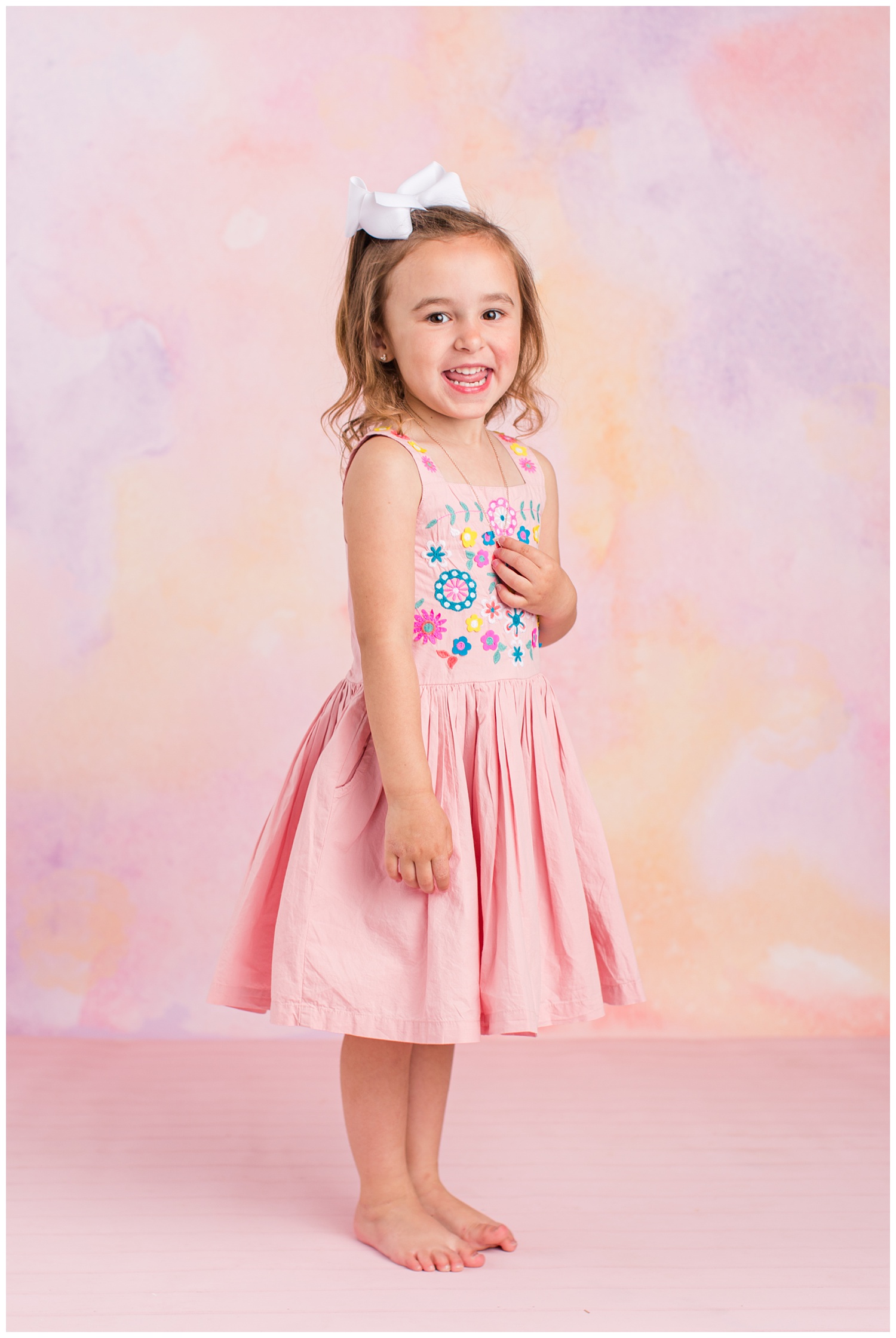 Four year old Sadie standing and smiling wearing a pink dress with floral embroidery on the bodice. 