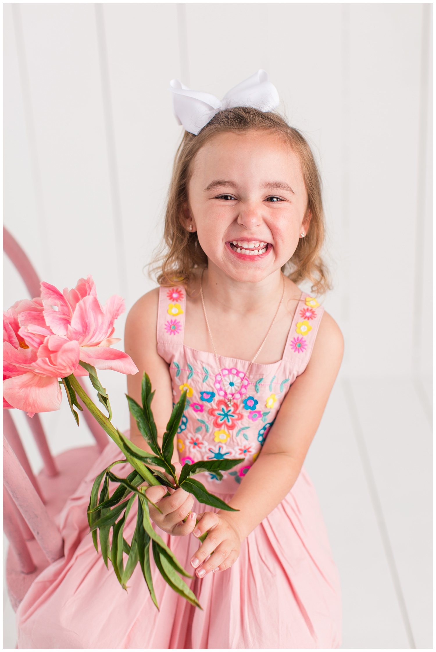 Four year old Sadie laughing while sitting on a pink chair wearing a pink dress with floral embroidered bodice and holding a pink peony stem.