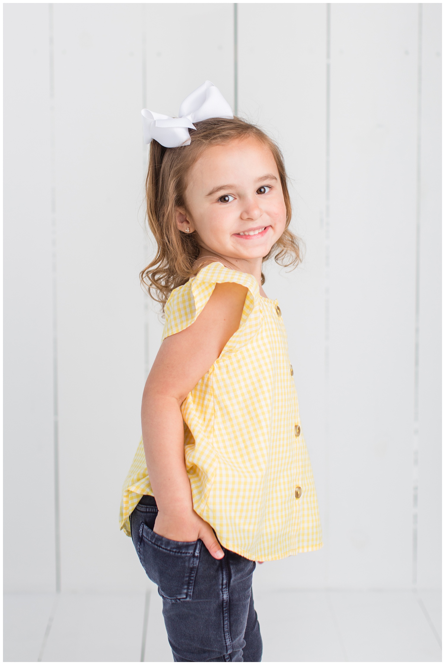 4 Year old Sadie wearing a yellow gingham top posing with her hand in her back pocket on a white wood background.
