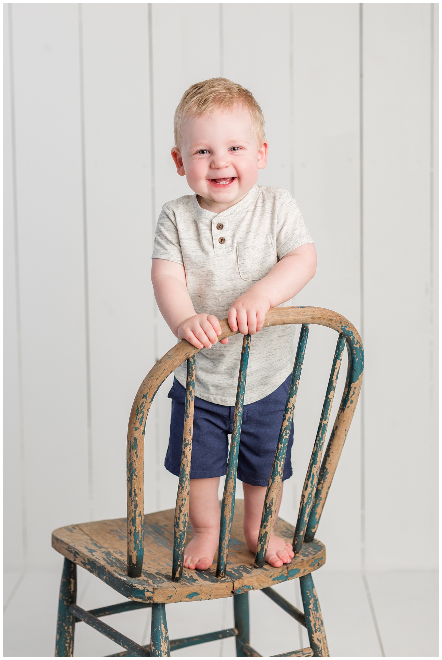Baby Carter standing on a rustic navy chair wearing a heather gray shirt and navy shorts smiling for his first birthday photo | CB Studio
