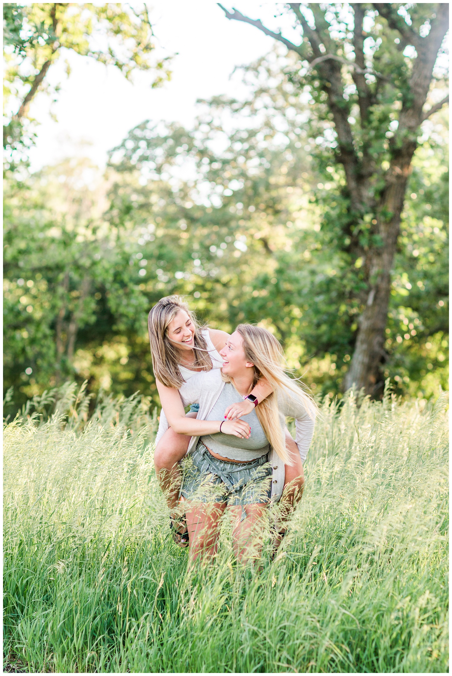Senior girls best friends BFFs piggy back while laughing in a grassy field on a rural Iowa farm | CB Studio