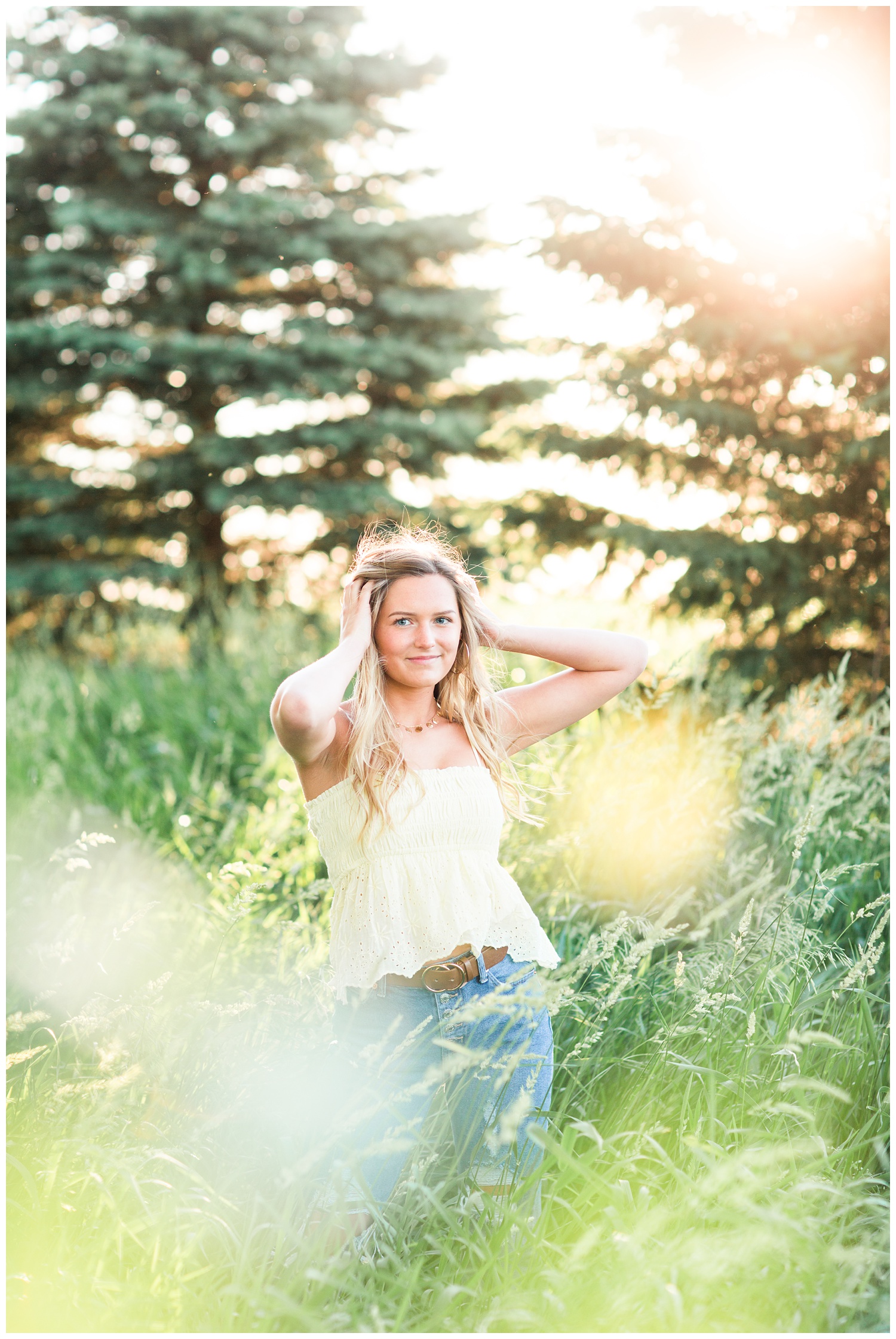 Senior girl wearing a yellow eyelet tank top standing in a grassy field with her hands in her hair. | CB Studio