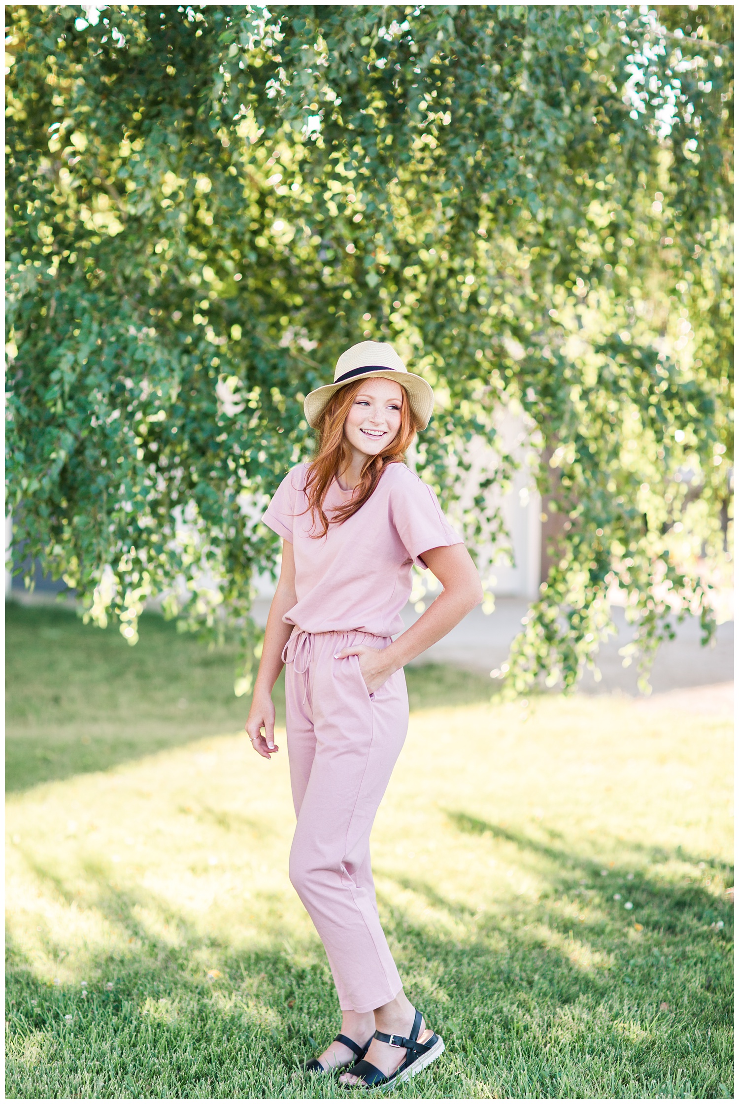 Senior girl wearing a dusty pink jumpsuit and a straw hat looking over her shoulder surrounded by birch tree branches in West Bend, Iowa | CB Studio 