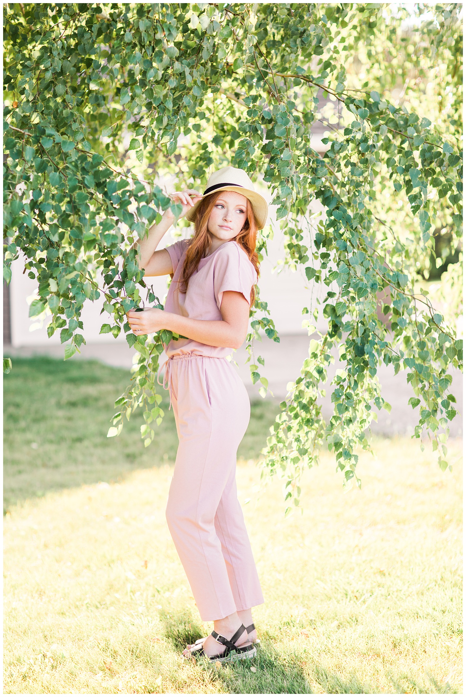 Senior girl wearing a dusty pink jumpsuit and a straw hat looking over her shoulder surrounded by birch tree branches in West Bend, Iowa | CB Studio 