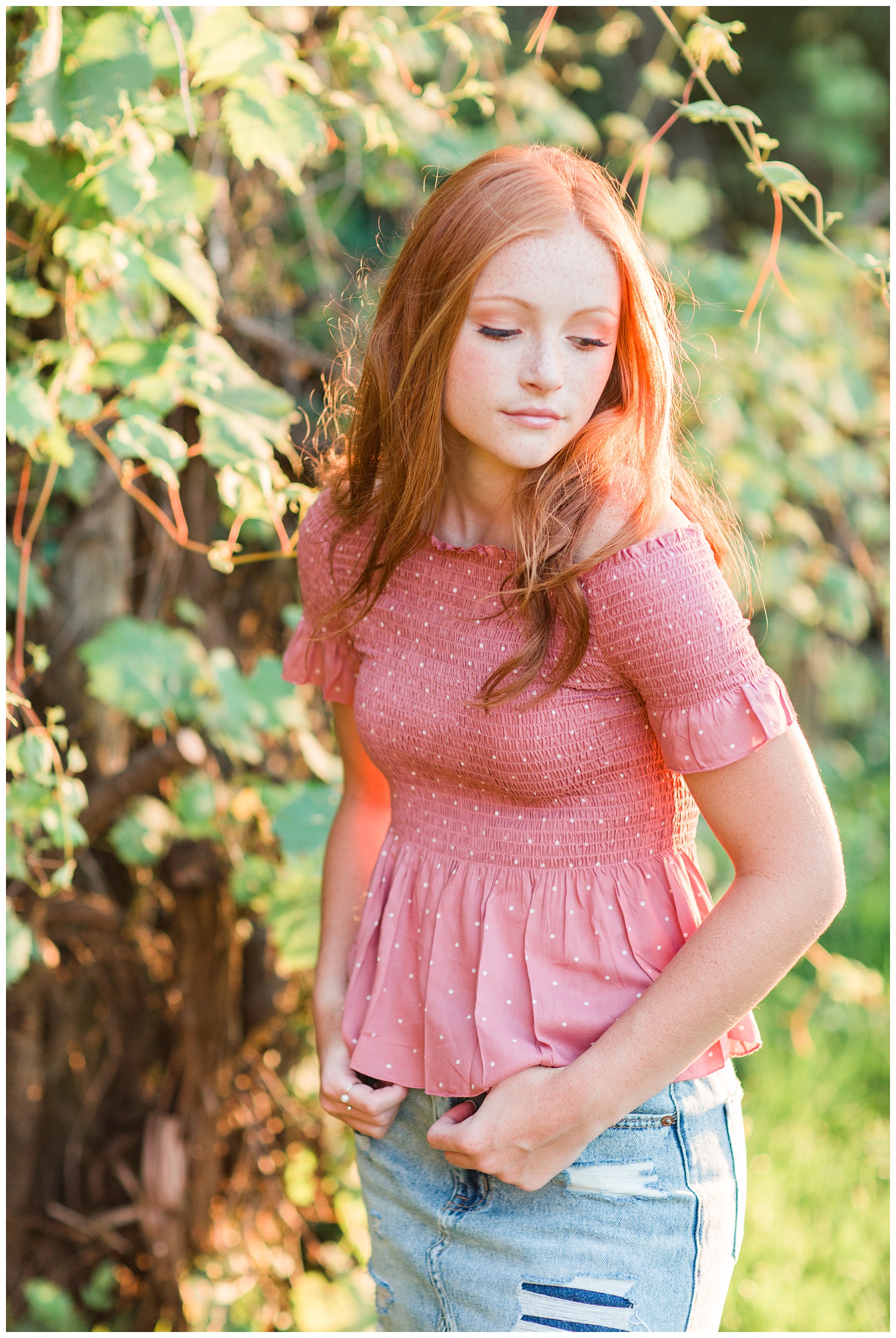 Senior girl wearing a pink, smocked, off the should top looking over her shoulder during golden hour in West Bend, Iowa | CB Studio