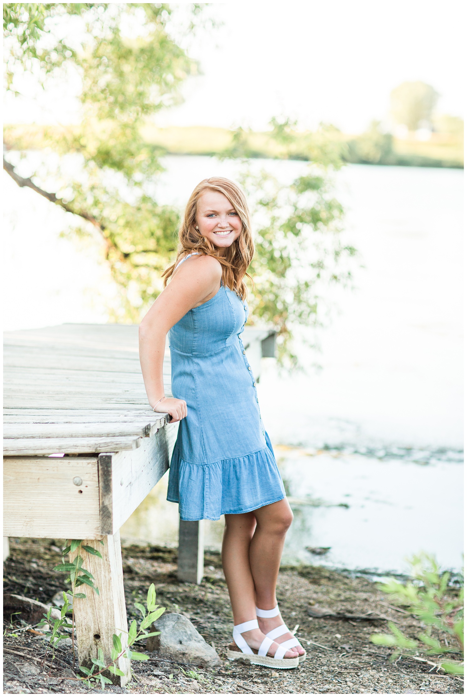 Senior girl wearing a denim dress standing next to a rustic wooden dock at Smith Lake, Algona, Iowa | CB Studio
