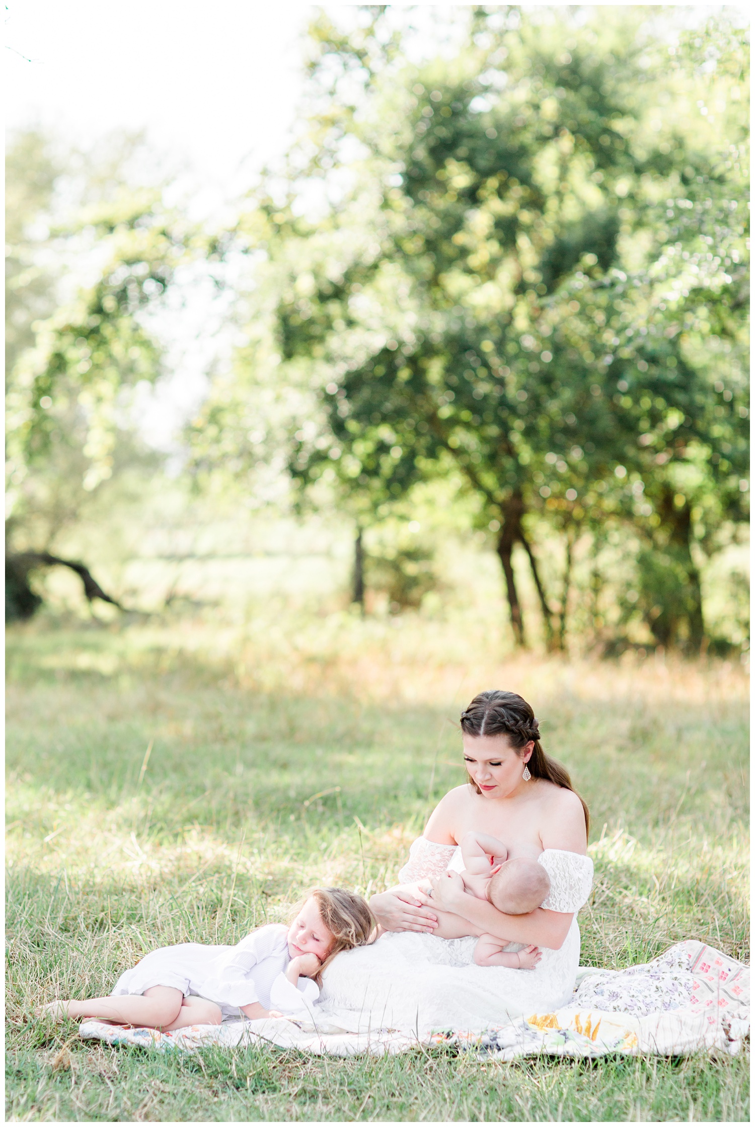 Beautiful mama wearing a white lacy dress sits in a glowy, grassy field breastfeeding her baby boy while her toddler girl rest upon her legs | CB Studio