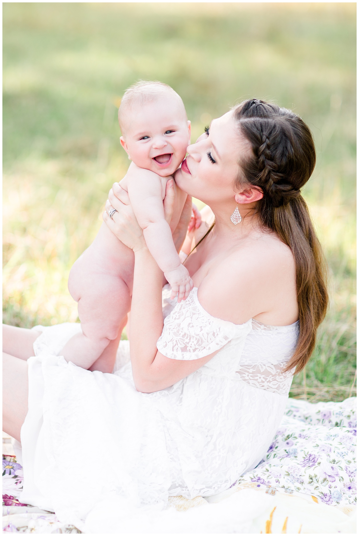 Beautiful mama wearing a white lacy dress sits in a glowy, grassy field snuggling her smiling baby boy | CB Studio