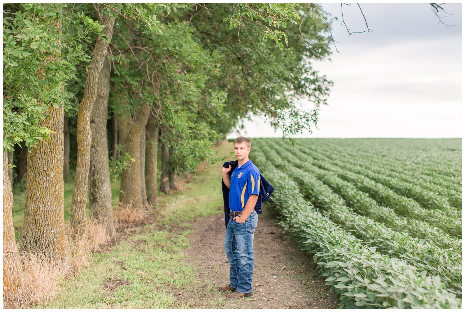 Senior boy wearing an FFA shirt stands next to a bean field holding his FFA jacket on a rural farm in Iowa | CB Studio