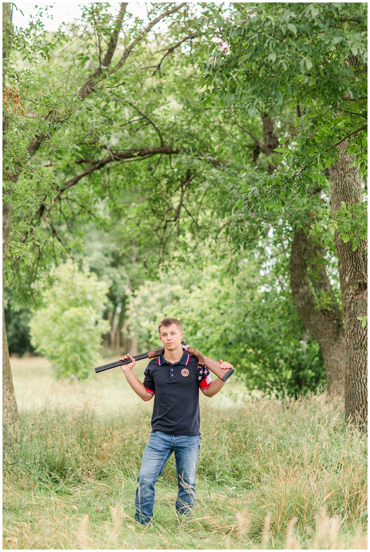 Senior boy wearing trapshooting gear carrying a shotgun around his neck in a grassy field surrounded by trees a on rural Iowa farm | CB Studio