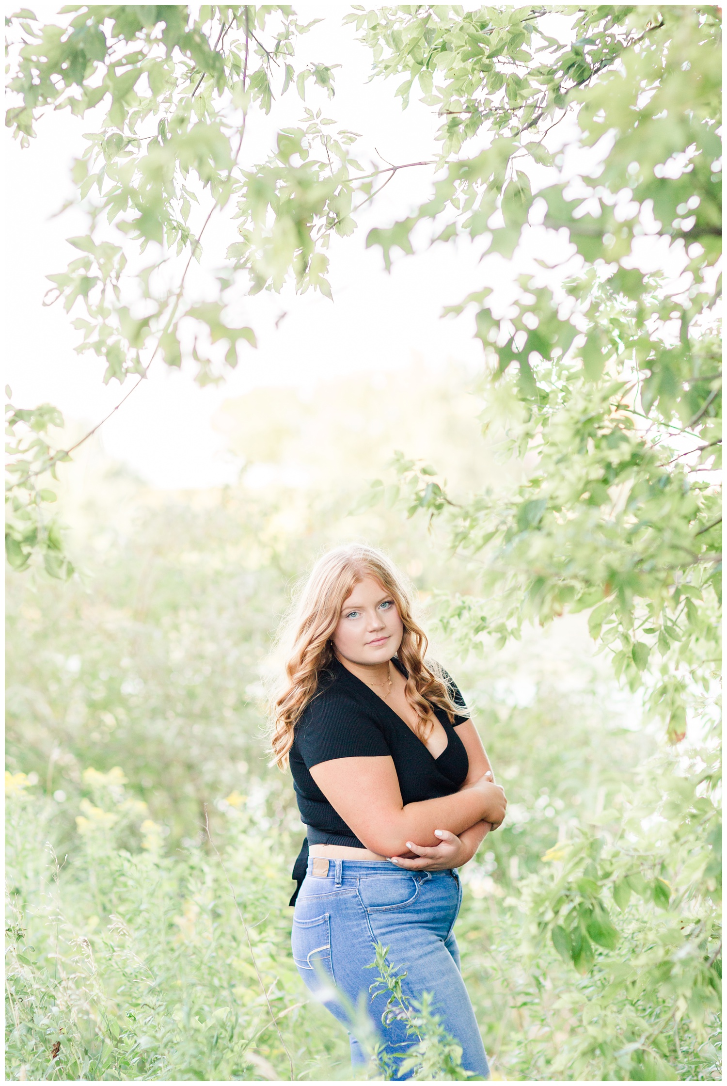 Senior girl wearing a black crop top and high waisted jeans stands surrounded by branches of leaves at Water's Edge Nature Center in Algona, Iowa | CB Studio