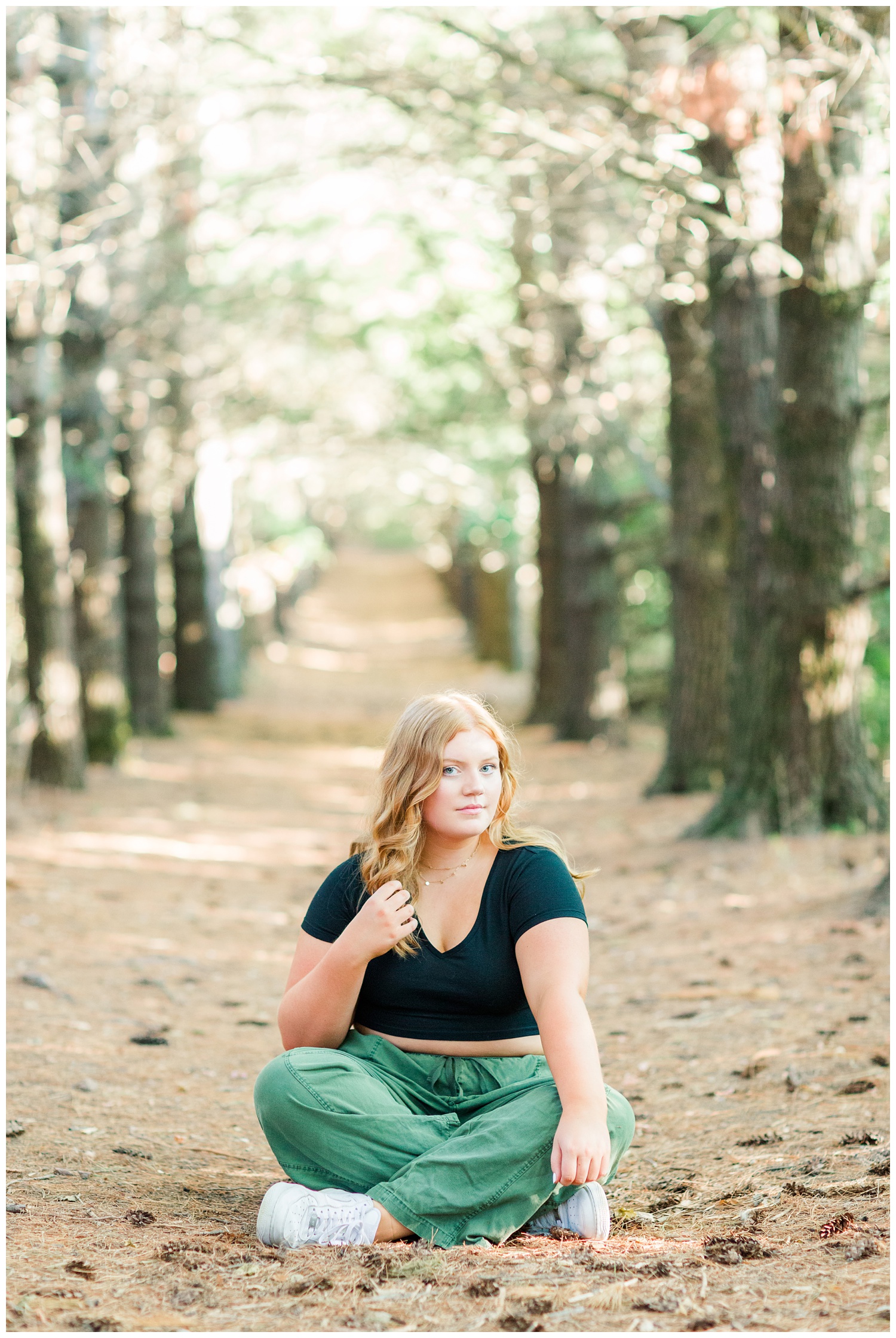 Senior girl wearing a black crop top and green cargo pants sitting beneath a row of pines at Water's Edge Nature Center in Algona, Iowa | CB Studio