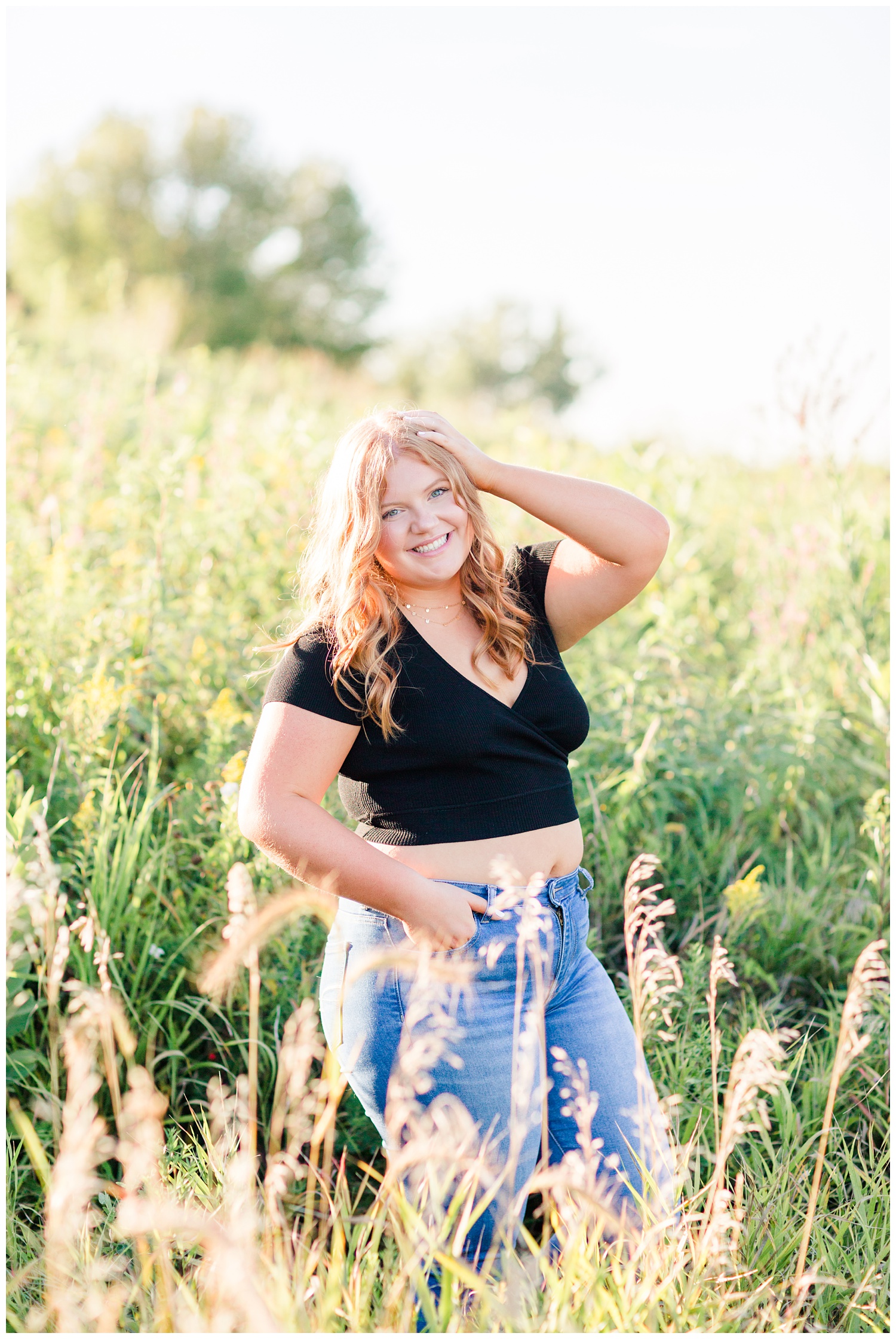 Senior girl wearing a black crop top and high waisted jeans stands in a grassy field at Water's Edge Nature Center in Algona, Iowa | CB Studio