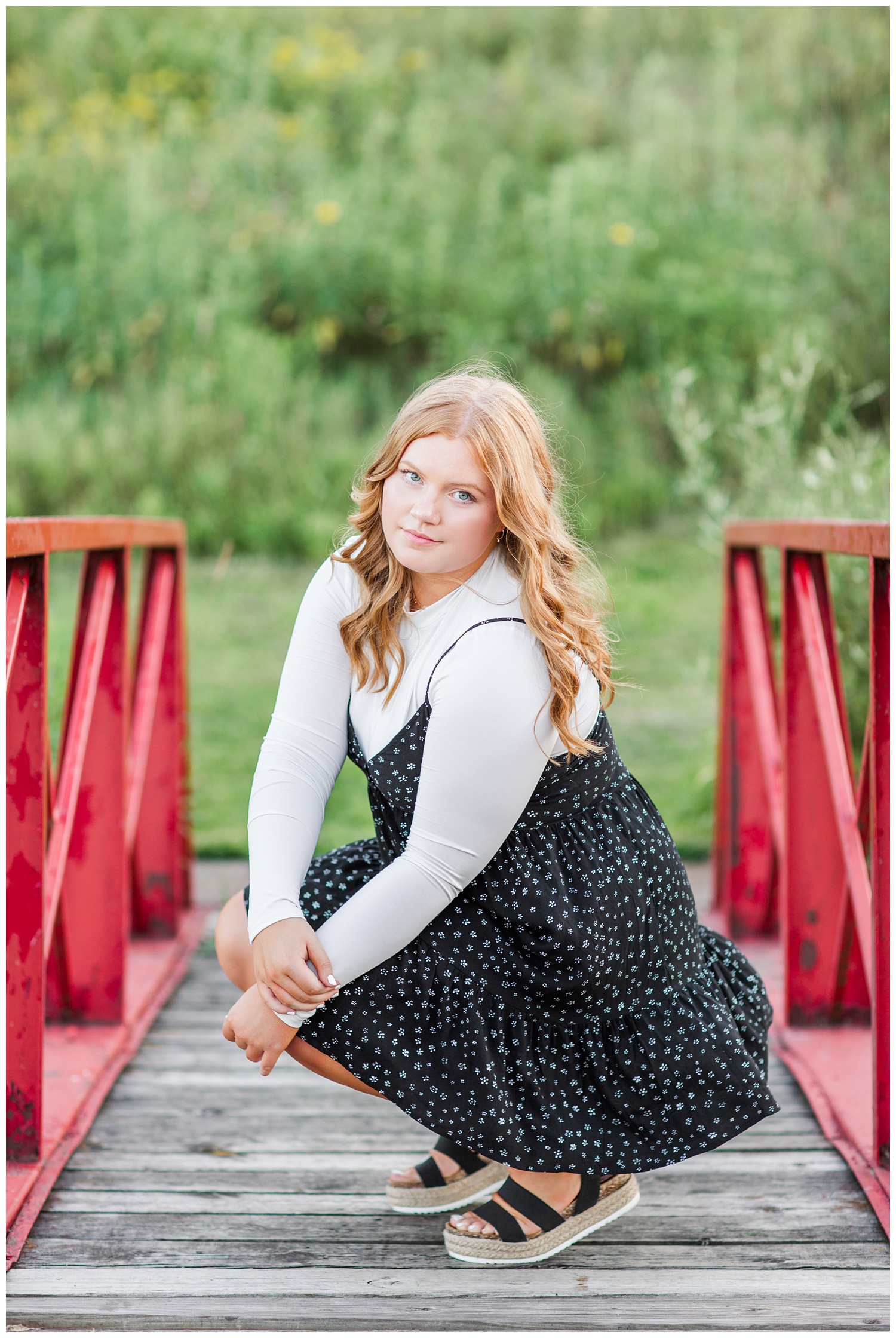 Senior girl wearing a black floral dress sits on a red bridge at Water's Edge Nature Center in Algona, Iowa | CB Studio
