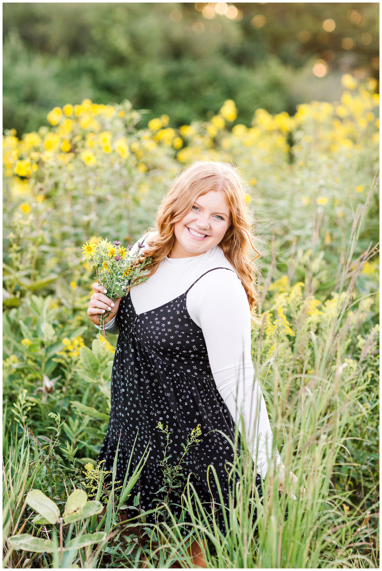 Senior girl wearing a black floral dress stands in a yellow flower field holding a flower bouquet at Water's Edge Nature Center in Algona, Iowa | CB Studio