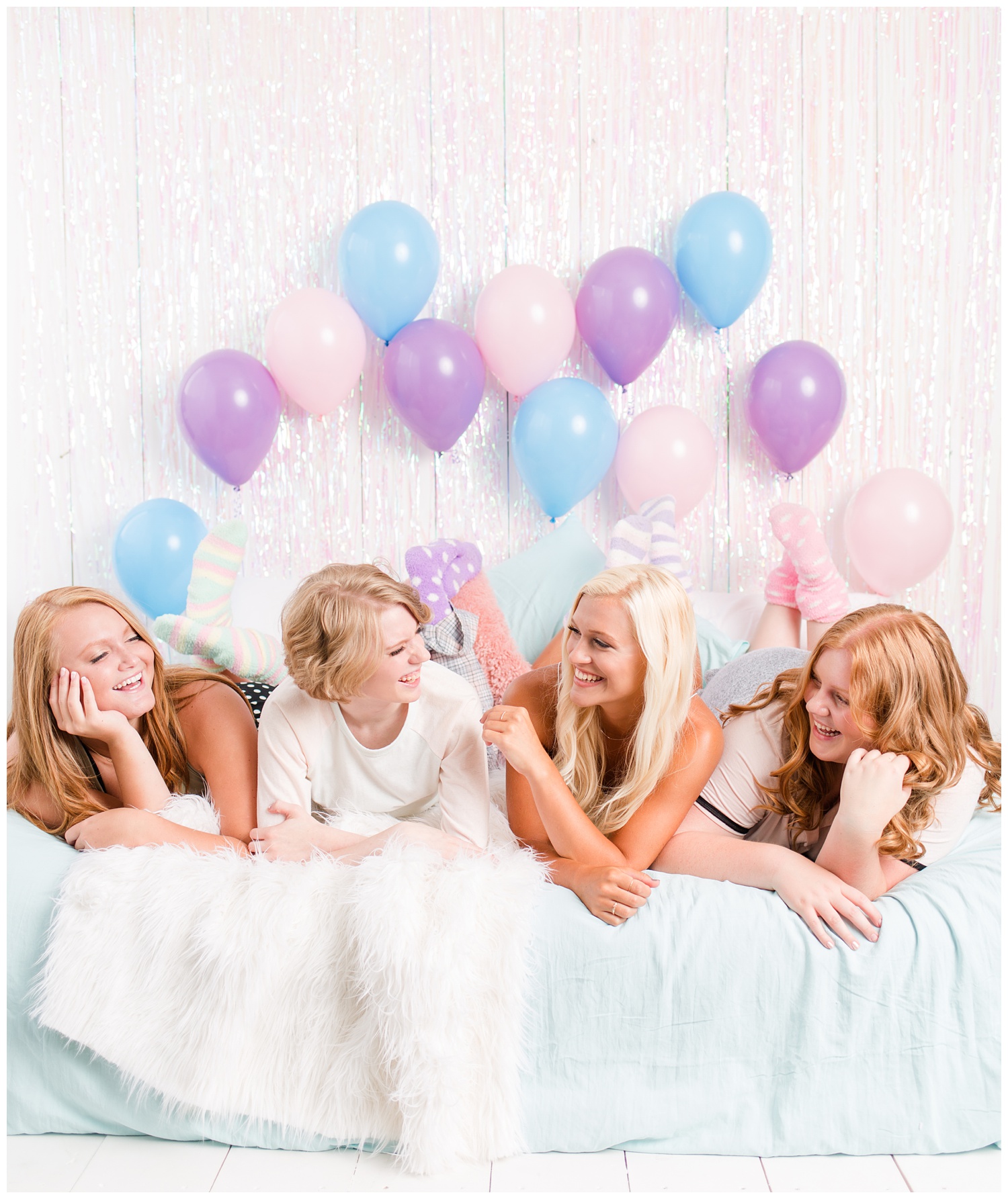 Senior girls laying on a bed and laughing during a senior sleepover-styled photoshoot with iridescent streamers and balloons. | CB Studio