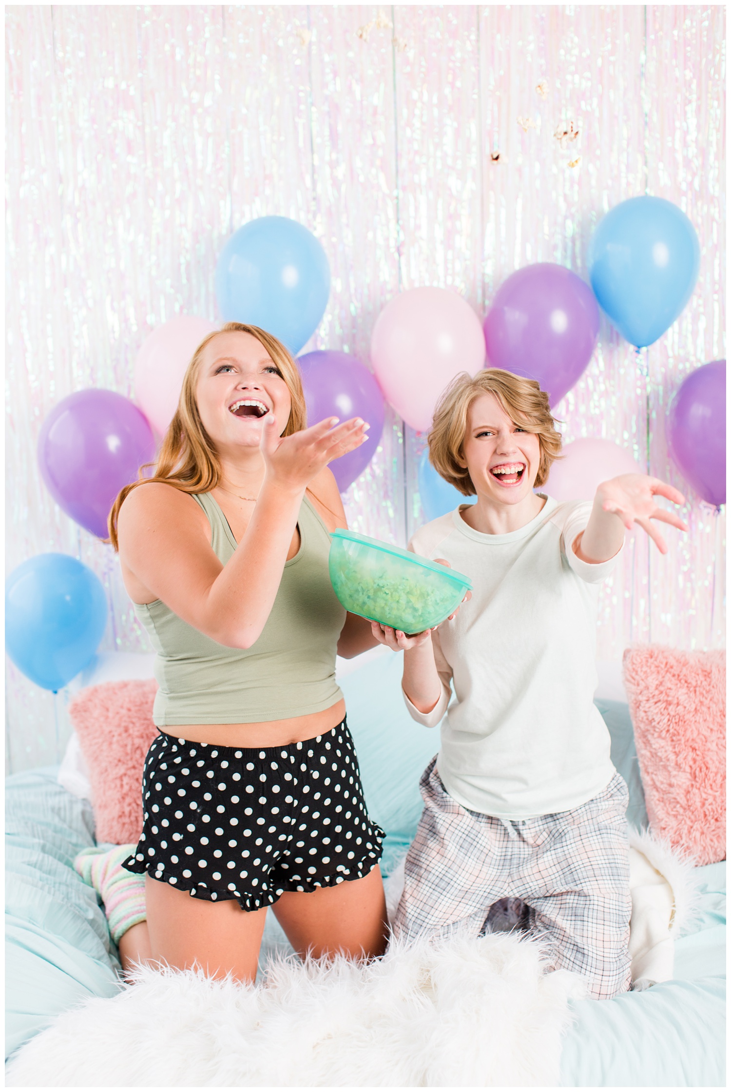 Senior girls throwing popcorn and laughing during a senior sleepover-styled photoshoot with iridescent streamers and balloons. | CB Studio