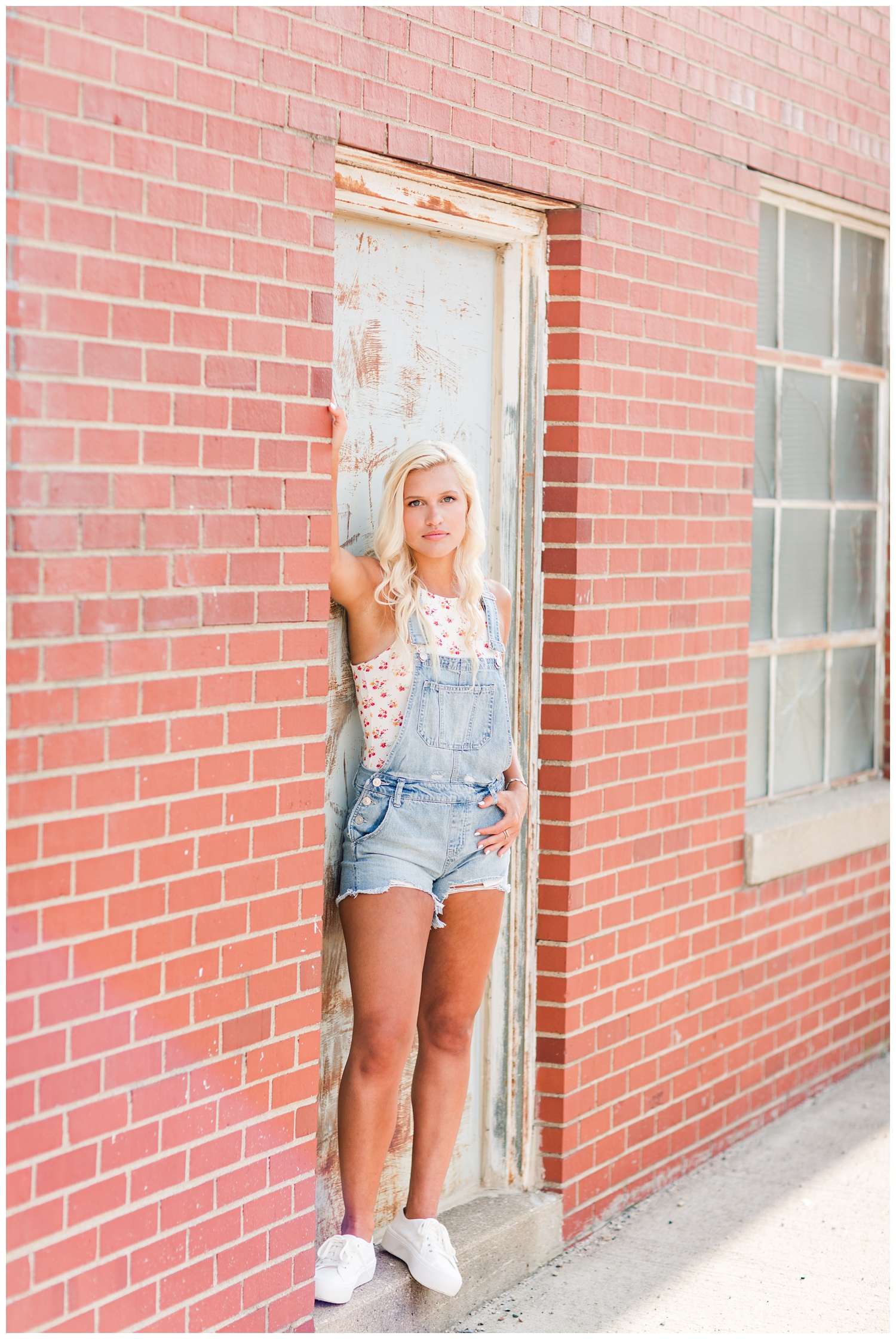Senior girl wearing jean overalls leans against a rusty wall in downtown Algona, Iowa | CB Studio