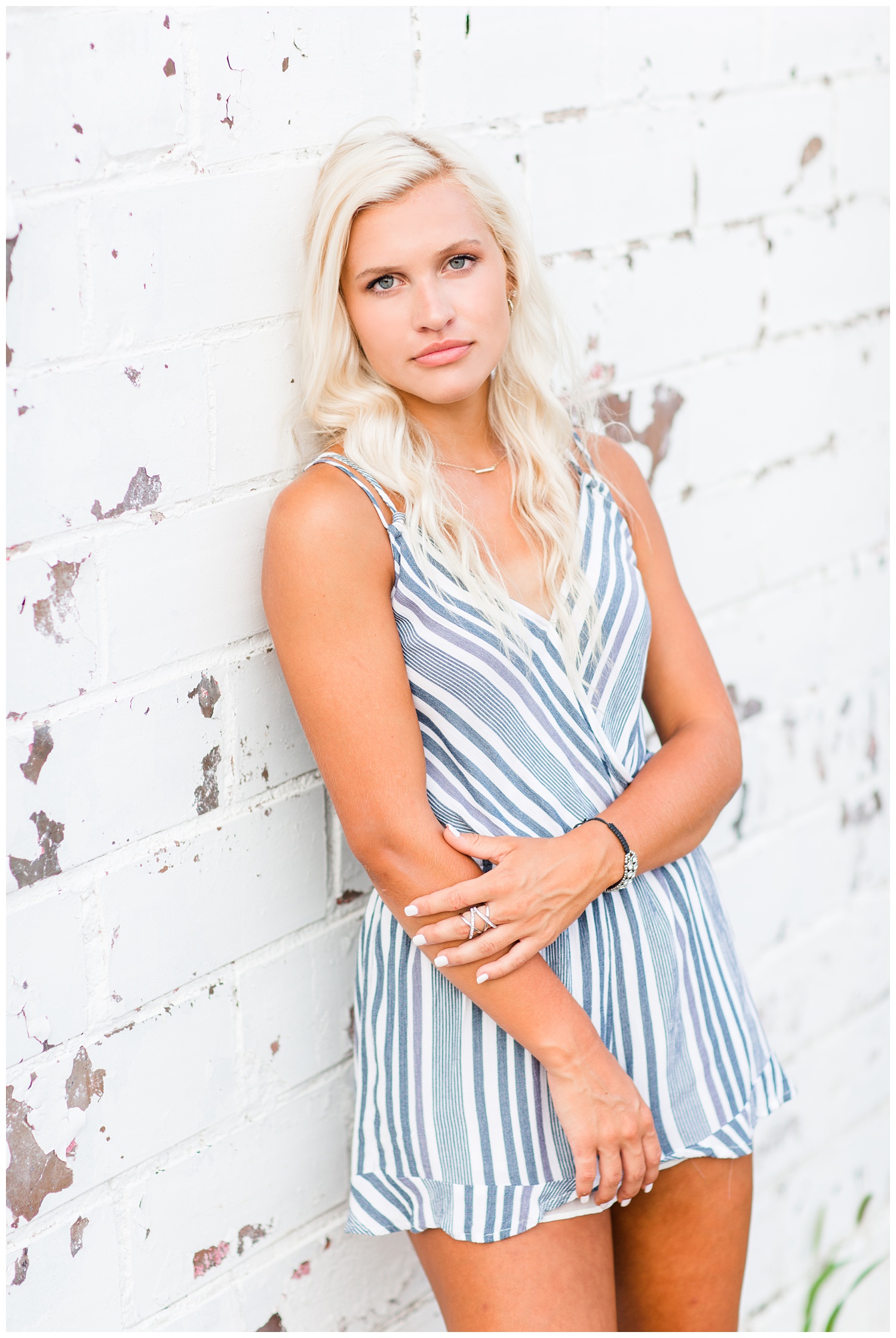 Senior girl wearing a white and blue romper leans against a rustic, white washed brick wall in downtown Algona, Iowa | CB Studio