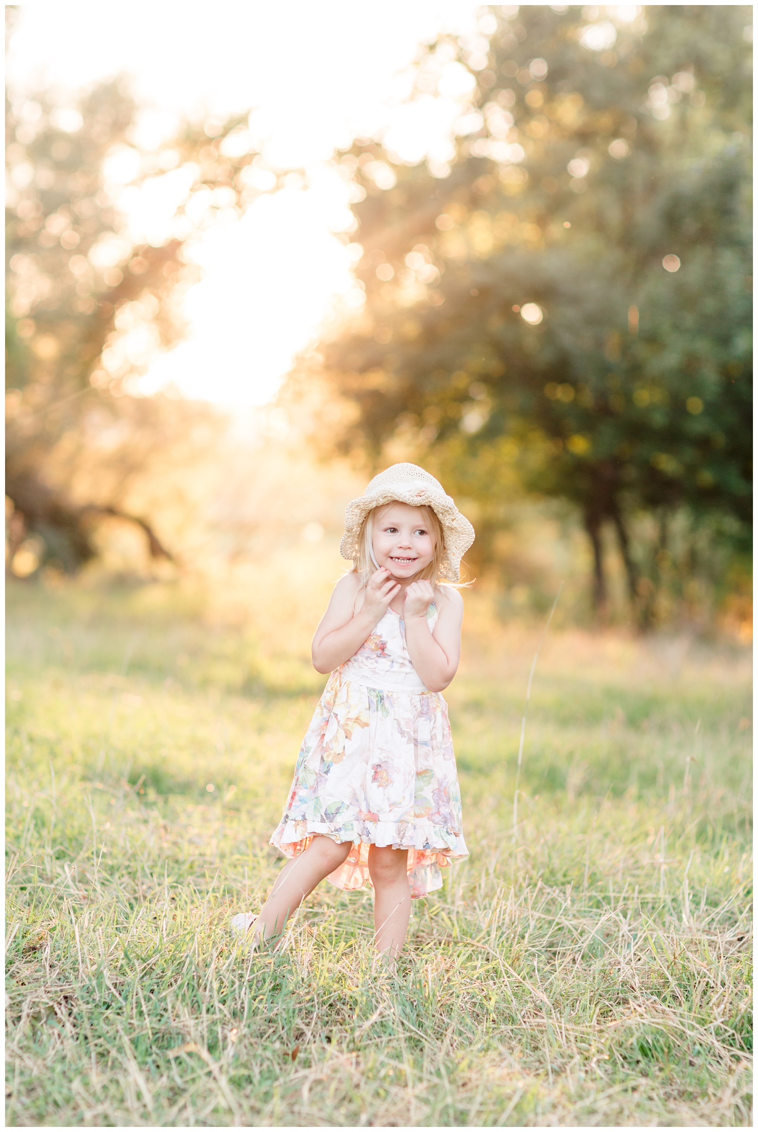 Little toddler girl wearing a floral dress and a straw hat dancing in a pasture during golden hour | CB Studio