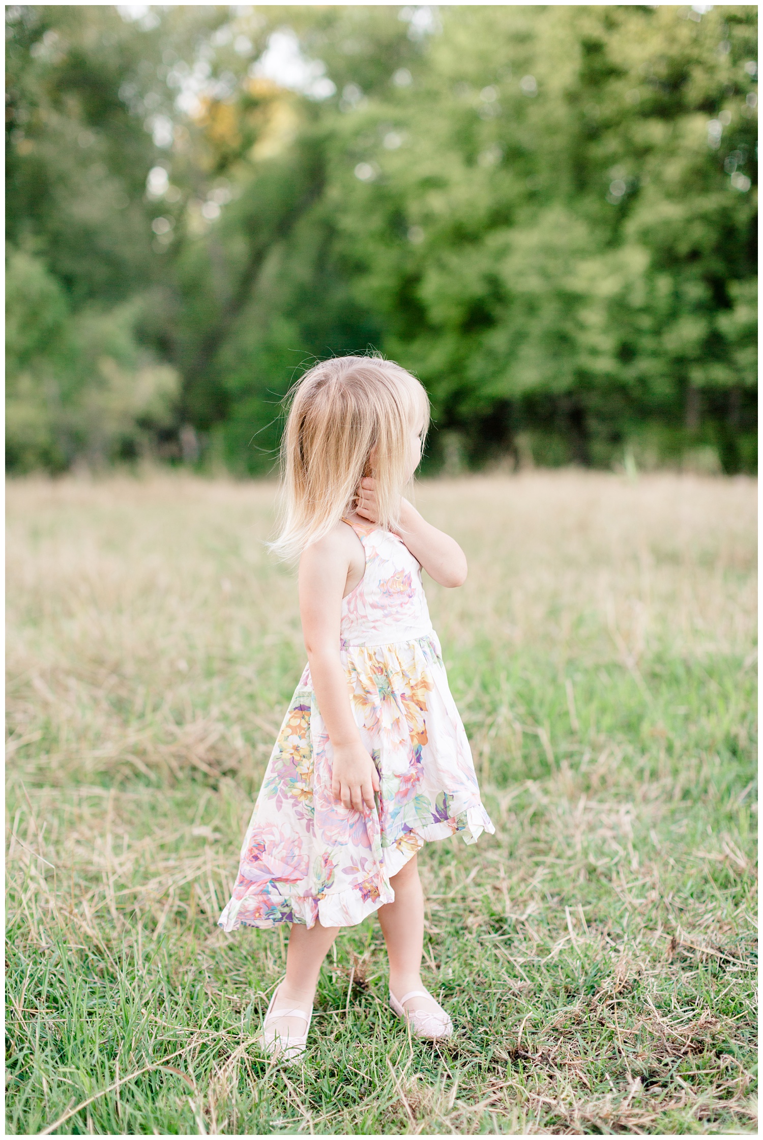 Little toddler girl wearing a floral dress looks off in the distance in a pasture during golden hour | CB Studio