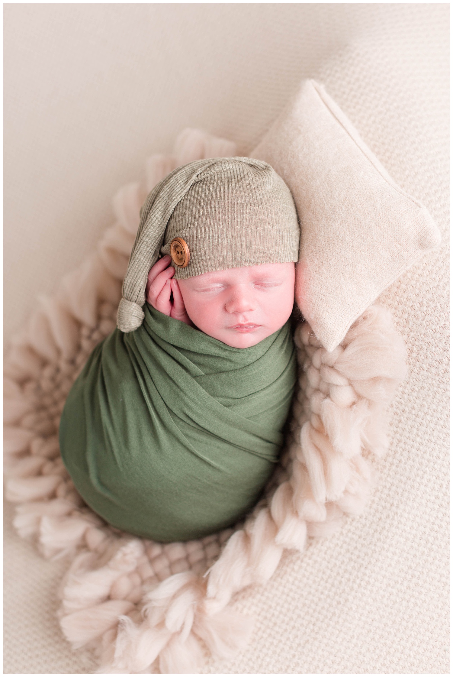 Newborn baby boy wrapped in a green swaddle wearing a green night cap with his hands gently folded along his cheek. | CB Studio
