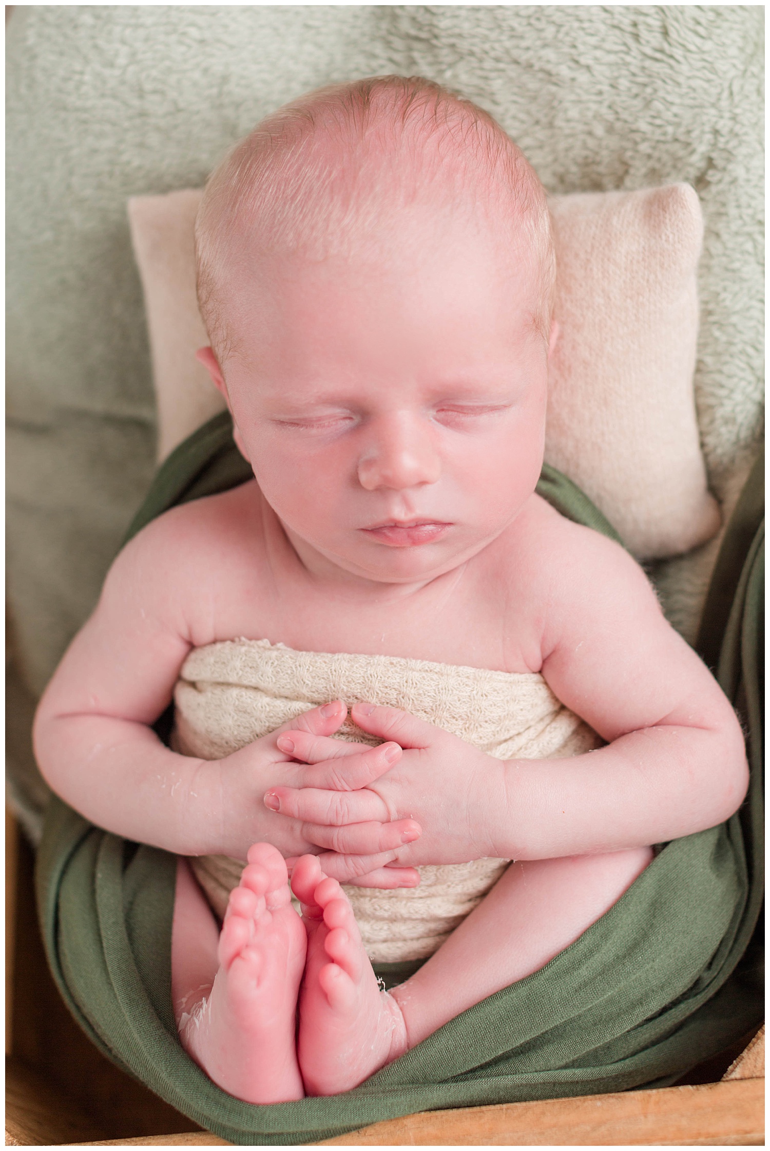 Newborn baby boy wrapped in nude and green swaddle with his hands and feet gently folded in his lap. | CB Studio