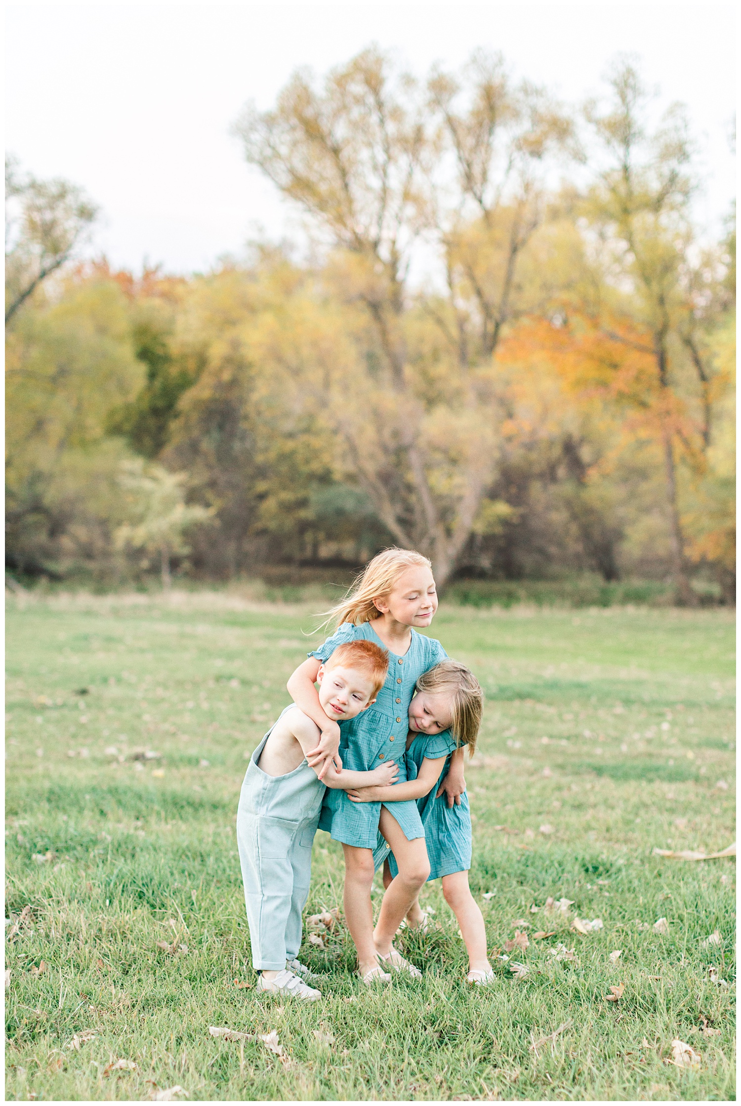 Three children dressed in vintage teal embrace each other in a grassy field looking onward | CB Studio